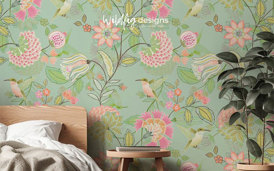 Whimsical floral wallpaper featuring pink and yellow flowers on a soft green background, used in a cosy bedroom setting. The room includes a beige throw blanket and a potted plant, enhancing the inviting atmosphere.