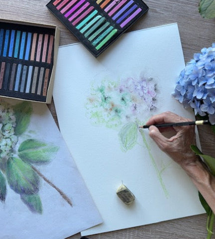 Wildfig designer Sam Drummond drawing flowers with pastels on a piece of paper, surrounded by art supplies.