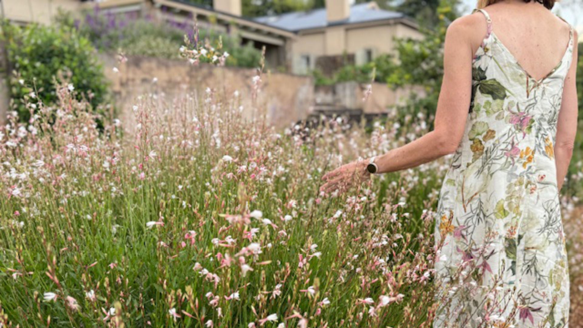 Woman in a floral dress standing in a field of tall grass and flowers with a house in the background.