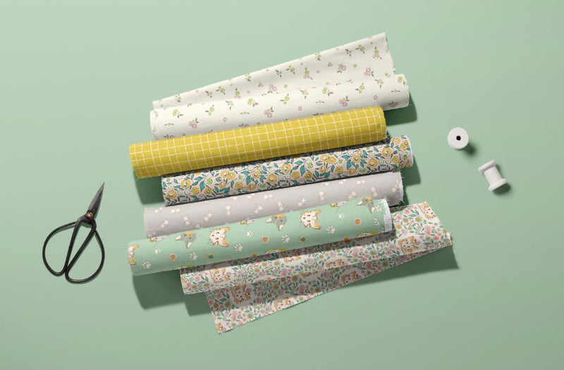 Rolls of floral and checked fabric on a green background with scissors and spools of thread. Suitable for kids apparel and nursery décor.